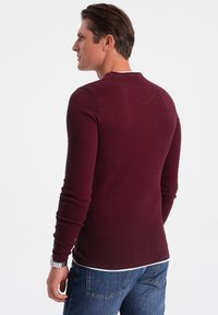 Burgundy textured long-sleeve shirt with a round neckline, paired with blue jeans, showing a fitted design and casual style.