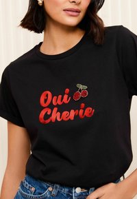 Must t-särk mustal taustal, millel on julge punane kiri "Oui Cherie", kaunistatud punase ja rohelise ehted-kirsi piniga. Lühikesed varrukad, ümmargune kaelus.