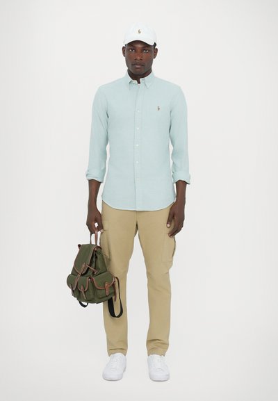 Chemise bleu clair à manches longues, pantalon cargo beige, baskets blanches, sac à dos vert avec des détails marron, et une casquette blanche avec un logo.