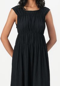 Black, sleeveless dress with vertical pleats, elastic waistband, and a round neckline, featuring a smooth texture and relaxed fit.