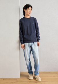 Sweatshirt azul-marinho de gola redonda com punhos canelados, combinado com jeans desbotados de um azul claro e sapatilhas bege, de pé contra uma parede cinza.
