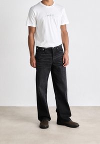 Camiseta blanca con el logo de "Calvin Klein Jeans", combinada con jeans negros holgados y zapatos marrones sobre un fondo neutro.