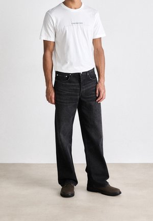 Witte T-shirt met het logo "Calvin Klein Jeans", gecombineerd met losse zwarte jeans en bruine schoenen op een neutrale achtergrond.