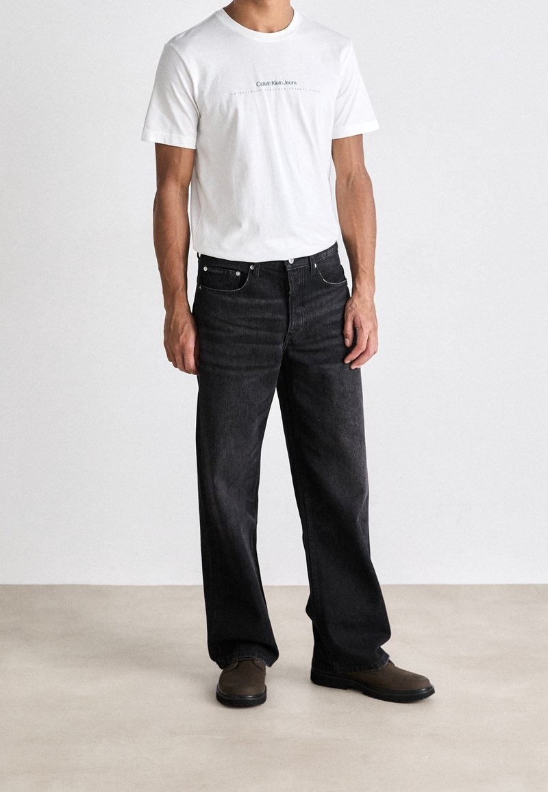 Camiseta blanca con el logo de "Calvin Klein Jeans", combinada con jeans negros holgados y zapatos marrones sobre un fondo neutro.