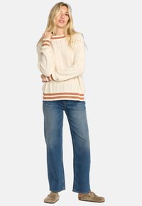 Pull en maille côtelée crème, avec des accents rayés marron aux manches et à l'ourlet. Associé à un jean en denim bleu à jambe droite et des chaussures beiges.