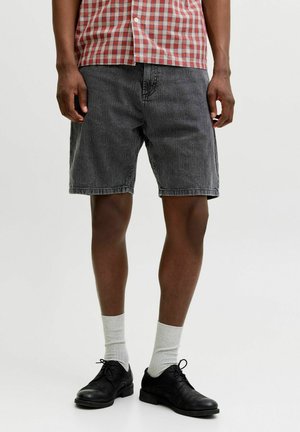 Mann trägt schwarze Jeans-Shorts, ein rot-weiß kariertes Hemd, weiße Sportsocken und schwarze Schnürschuhe, steht vor einem schlichten Hintergrund.