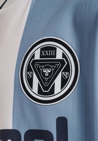Lichtblauw shirt met een zwart rond embleem met een geometrisch ontwerp en het cijfer "XXIII" erop. De stof lijkt glad met een subtiele textuur.