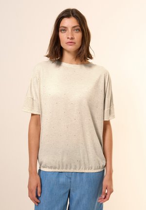 Helles graues T-Shirt mit kurzen Ärmeln und lockerer Passform, das mit kleinen blauen Punkten verziert ist, einen runden Ausschnitt hat und einen elastischen Saum. Kombiniert mit blauen Hosen.