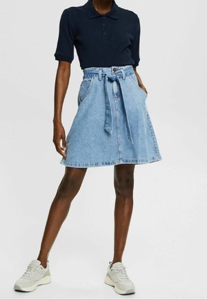 Femme portant un polo bleu marine à manches courtes, une jupe en jean bleu clair taille A avec ceinture, et des baskets blanches, debout les mains dans les poches.