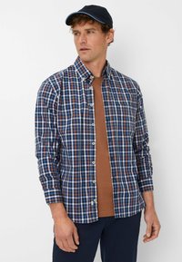 Langarm, knoopshirt met een blauw, bruin en wit geruit patroon. Heeft een puntige kraag en omgeslagen manchetten. Bruine T-shirt eronder.