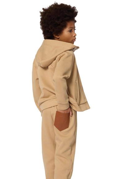 Beige Kapuzenpullover und Jogginghosen-Set, weicher Stoff, Kängurutasche, Kapuze mit innerem Muster und eine Lederpatch-Tasche auf der Hose.