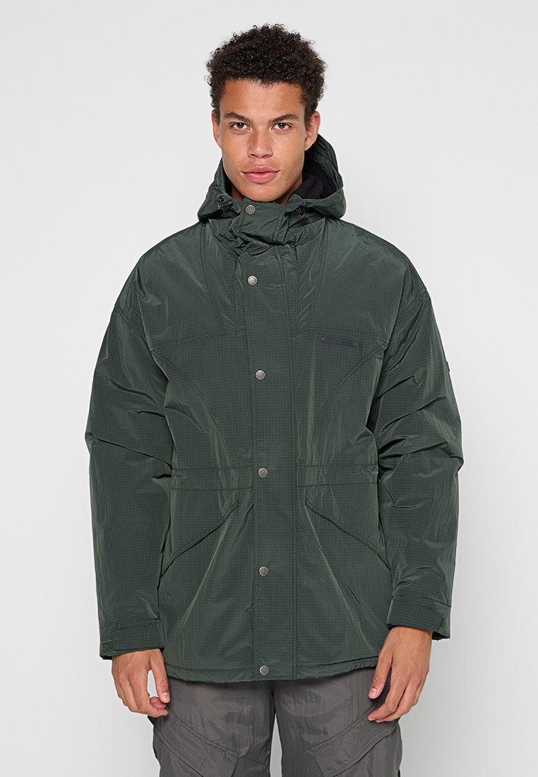 Mężczyzna w ciemnozielonej zimowej kurtce z kapturem, zapinanej na napy, oraz szarych spodniach cargo, stojący na jednolitym jasnym tle.