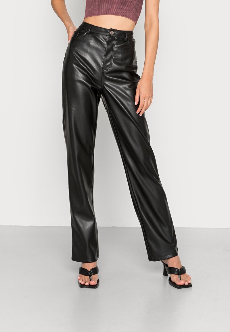 Femme portant un pantalon droit en similicuir noir taille haute et des sandales à talons noires, debout avec une main sur la hanche.
