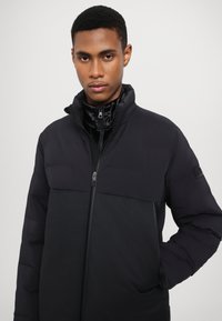 Black puffer jacket with a zip closure, featuring a solid top section and textured lower section. Smooth fabric with a matte finish.