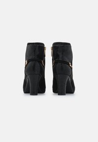 Par de botas de tobillo negras texturizadas con tacones cuadrados, cremalleras laterales y correas trenzadas decorativas con acentos de hebilla en tono dorado, vistas desde la parte posterior.