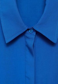 Camicia blu a colletto in tessuto liscio, con chiusura a pulsante e un colletto pulito e nitido. Dettagli cuciti in blu abbinato.