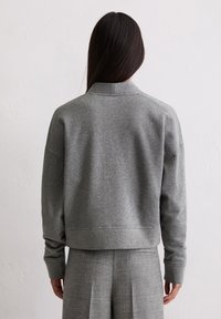 Sweatshirt gris coupé avec un col montant et des poignets côtelés. Le tissu est lisse avec une texture douce, offrant une coupe décontractée et des épaules tombantes.