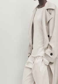Trench-coat beige avec des boutons noirs, porté sur un haut en maille beige clair et un pantalon ample, affichant une texture lisse.