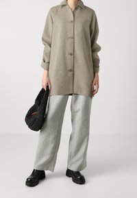 Vrouw met een beige getailleerde jas met knopen, lichtgrijze wijde broekspijpen, zwarte stevige laarzen, die een zwarte gebreide handtas vasthoudt tegen een effen achtergrond.