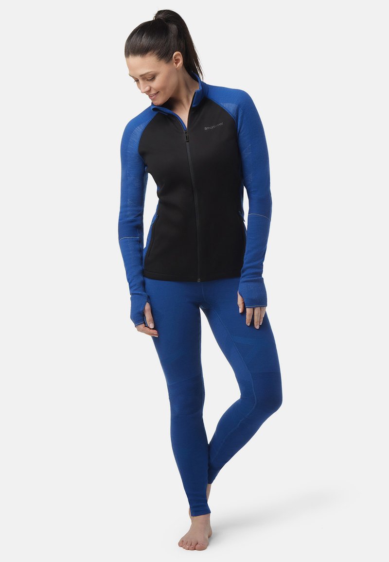 Zwarte zip-up sportjack met blauwe mouwen, gecombineerd met blauwe legging. Kenmerken zijn duimgaten en textuurelementen voor extra comfort.