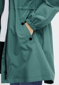 Jachetă impermeabilă verde cu manșete elastice, închidere cu fermoar și un buzunar lateral cu clapetă, având o textură netedă și un design casual.