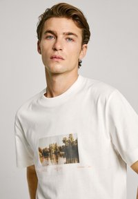 Balts kokvilnas T-krekls ar ainavas fotogrāfiju uz priekšpuses. Dizainā ietilpst brīvs piegriezums un ribota apkakle.