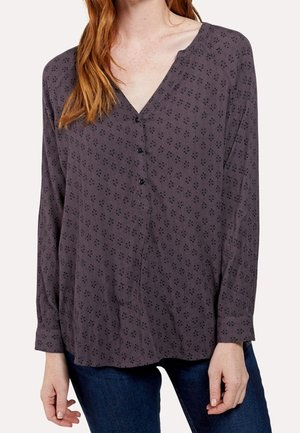 Mujer que lleva una blusa de manga larga de color gris oscuro con un sutil estampado floral, escote en V, tres botones, combinada con vaqueros azul oscuro.