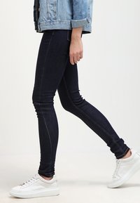 Jeans skinny de color azul oscuro con costuras en contraste, combinados con zapatillas blancas. Los jeans tienen un diseño ajustado y una cintura estándar.