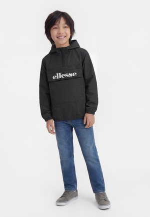 Bambino sorridente che indossa una giacca con cappuccio nera ellesse, jeans blu e scarpe da ginnastica grigie, in piedi davanti a uno sfondo bianco uniforme.