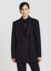 Blazer nero su misura con design avvolgente, dettaglio con laccio in vita, maniche lunghe e colletto strutturato. Tessuto dalla texture liscia.