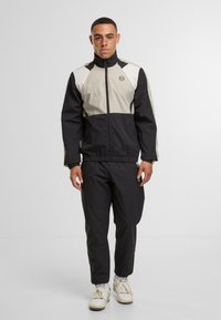 Giacca a vento nera e beige con colletto alto, zip frontale e polsini elasticizzati. Pantaloni neri e sneakers bianche.