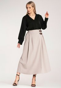 Czarna bluzka z długim rękawem w połączeniu z jasnobeżową, plisowaną maksisukienką, z dekoracyjnymi brązowymi klamrami w talii. Czarne sandały na obcasie.