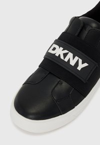 Baskets en cuir noir avec bride en textile arborant le logo "DKNY" blanc. Semelle en caoutchouc blanc et détails de couture. Texture lisse et bout arrondi.