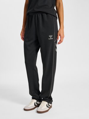 Osoba ubrana w czarne spodnie dresowe Hummel z bocznymi paskami oraz białe sneakersy z czarnymi wzorami w zygzak stoi na jednolitym tle.