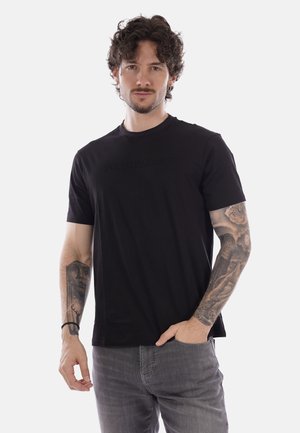 Uomo con capelli ricci che indossa una maglietta nera Armani Exchange e jeans grigi, mostrando tatuaggi dettagliati su entrambi gli avambracci.
