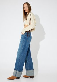 Top corto de punto crema con flecos laterales, combinado con jeans de mezclilla azul de pierna ancha que tienen un dobladillo de mezclilla y botas de tobillo de gamuza marrón.