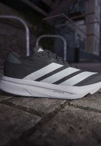 Μαύρο και λευκό παπούτσι τρεξίματος Adidas με τρεις λευκές ρίγες και σόλα Lightstrike Pro που ακουμπάει σε λιθόστρωτο.