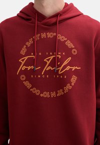 Felpa bordeaux con testo stampato in oro. Presenta un design rotondo con le coordinate e "Tom Tailor" in evidenza. Cappuccio regolabile con coulisse.
