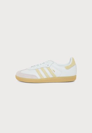 Λευκό αθλητικό παπούτσι Adidas Samba με μπεζ σουέτ λεπτομέρειες, τρεις υφασμάτινες ρίγες και λαστιχένια σόλα, απεικονίζεται από το πλάι σε λευκό φόντο.