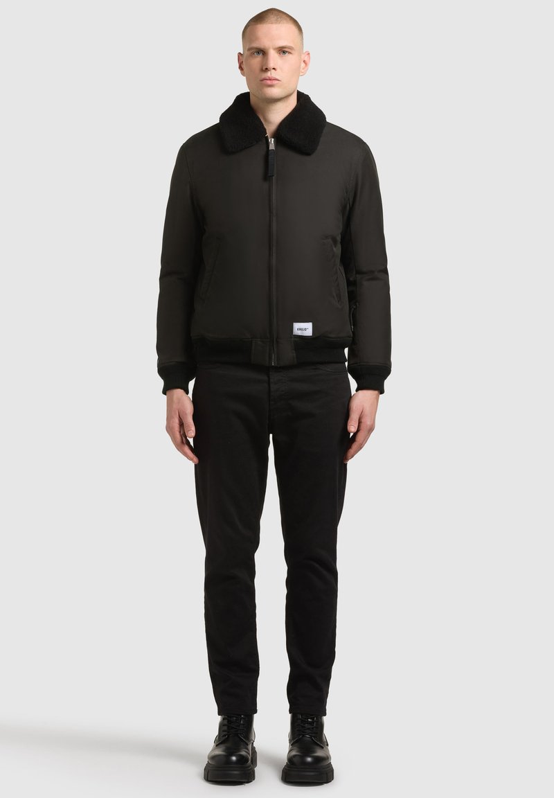 Czarna kurtka bomberka z miękkim kołnierzem wykończonym futerkiem, zamkiem błyskawicznym oraz ściągaczami przy rękawach, zestawiona z czarnymi spodniami i butami.