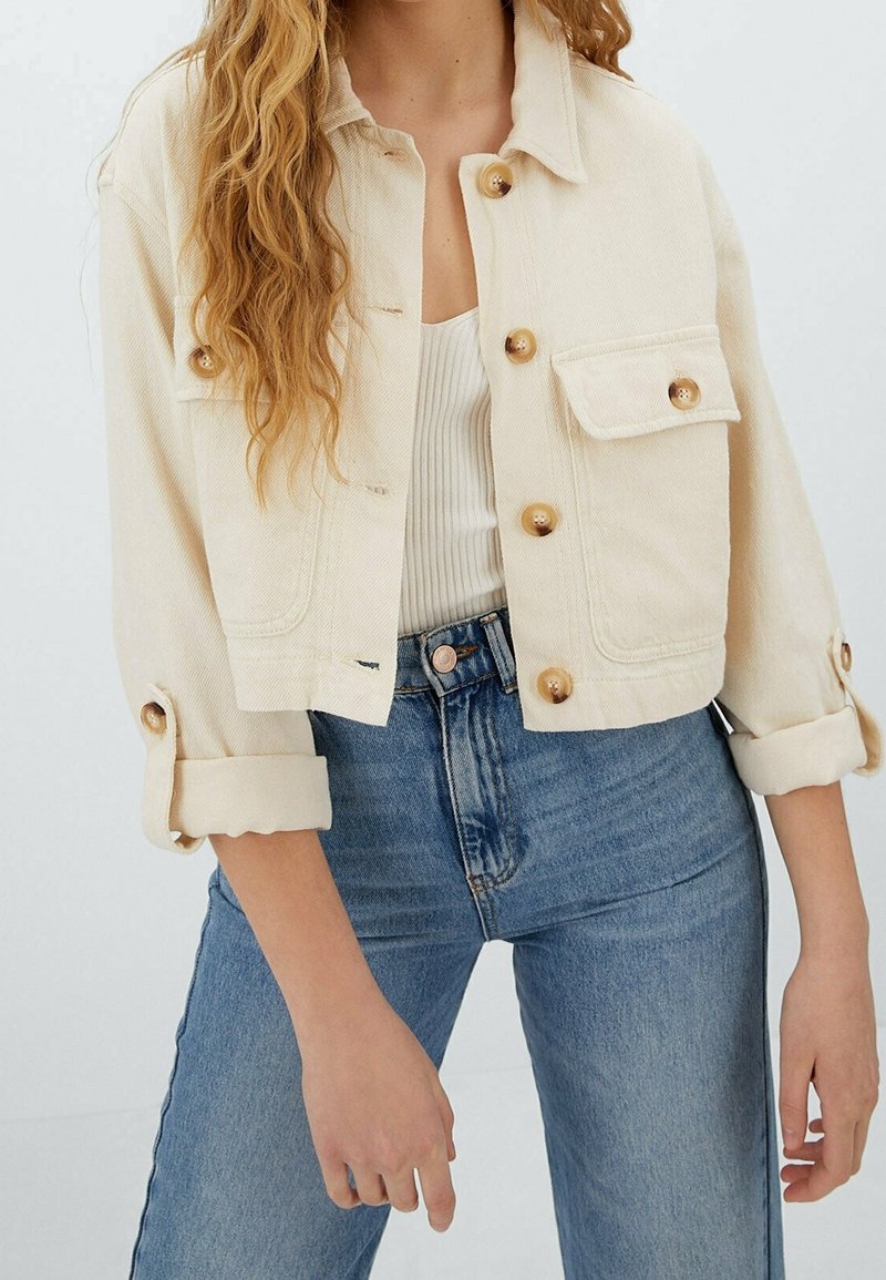 Kremowa, krótka kurtka dżinsowa z złotymi guzikami, kieszenią na piersi i podwiniętymi rękawami, noszona na białym, prążkowanym topie i niebieskich dżinsach.