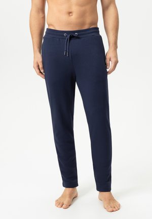 Mann trägt dunkelblaue Sweatpants mit Kordelzug und steht barfuß vor einem schlichten hellen Hintergrund.