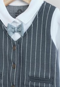 Szürke és fehér csíkos mellény fehér gallérral, denimszínű csokornyakkendővel és barna gombokkal, egy fából készült vállfán bemutatva.