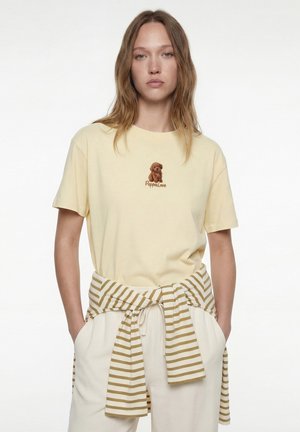 Mujer con una camiseta de color amarillo pálido con un pequeño gráfico de un perro marrón y texto "Peppulore", pantalones color crema y un suéter a rayas atado alrededor de su cintura.