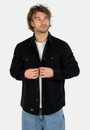 Black corduroy shirt with two chest pockets, button closure, and a relaxed fit. Worn over a white t-shirt and paired with light blue jeans.
