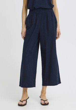 Nő sötétkék, bőszárú, rövidített nadrágot visel, amely gumis derékrészű, fekete magassarkú szandálban, és egy egyszerű világos háttér előtt áll.