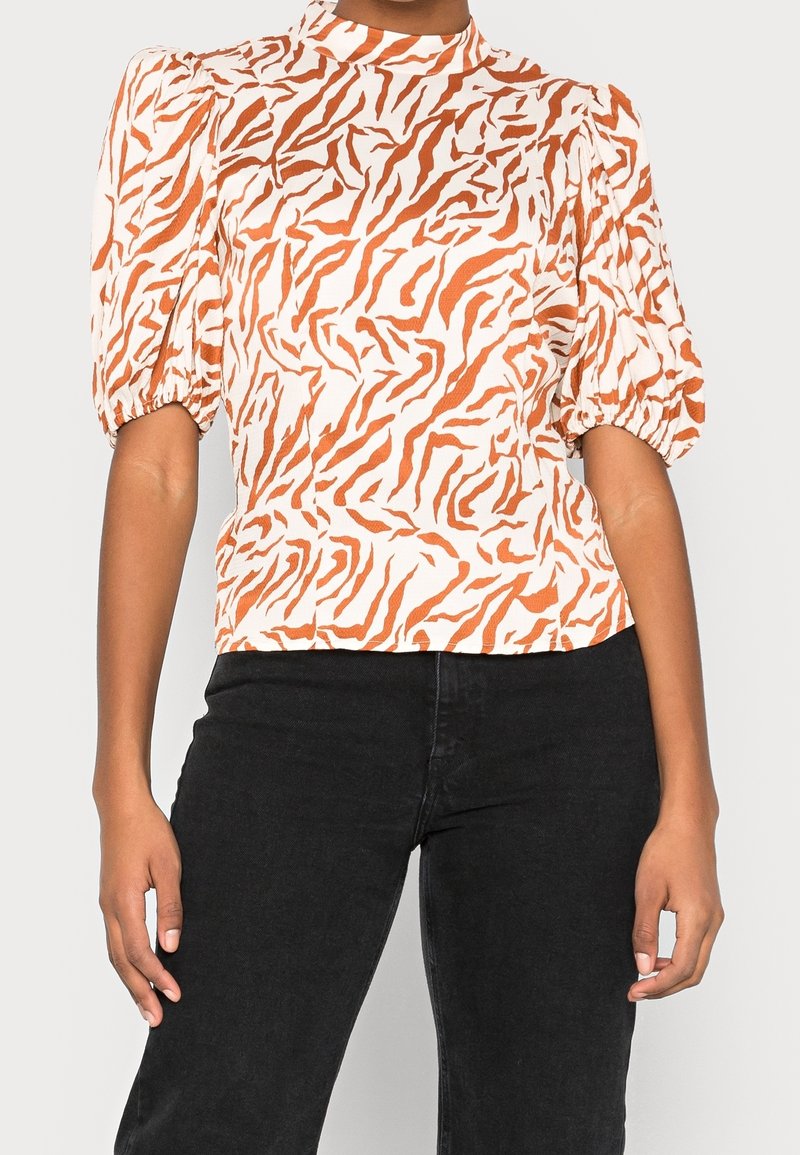 Blusa bianca con motivo a strisce di tigre arancioni, collo alto, maniche a sbuffo e silhouette aderente. Abbinata a jeans neri.