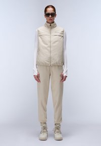 Gilet rembourré beige avec col montant sur un haut à manches longues blanc, associé à un jogging beige assorti et des bottes à lacets de couleur claire.