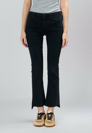 Osoba ubrana w czarne jeansy typu bootcut i beżowe trampki, stojąca na neutralnym szarym tle.