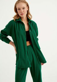 Grün, texturiertes Hemd mit Kragen, kombiniert mit passenden Hosen mit Gummizug in der Taille. Das Material hat ein weiches, geripptes Aussehen.